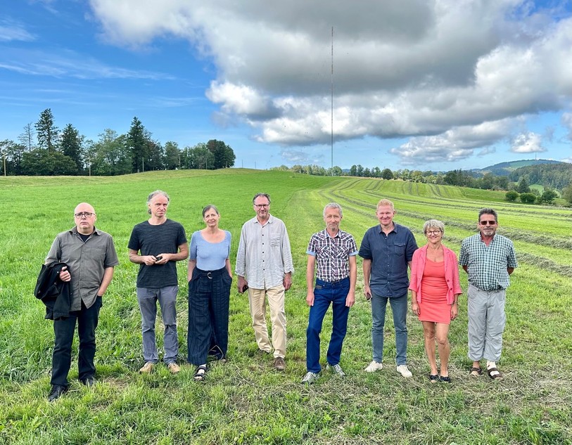 De gauche à droite : Romain Crelier, Philippe Queloz, Pascale Hoffmeyer, Herbert Kähli, Michel Fior, Marc Mertenat, Mimi Mertenat et Jean-Marc Baume posent devant un des mâts d'éoliennes érigés dans le cadre du projet éolien de la Haute-Borne.