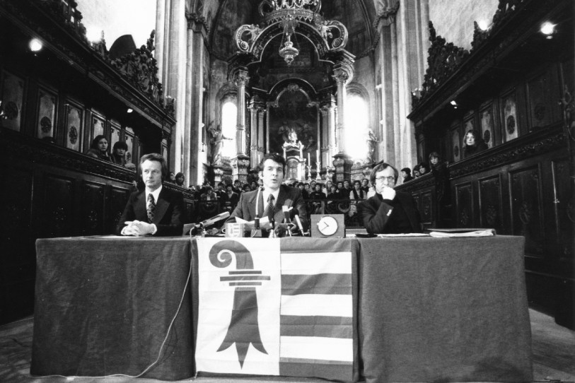 Roland Béguelin, François Lachat et Joseph Boinay (de gauche à droite) étaient à la tribune lors de la cérémonie d'acceptation de la Constitution jurassienne à la collégiale de Saint-Ursanne en février 1977. (Photo : Archives cantonales jurassiennes, Fonds Roland Béguelin (97 J RB 175), CC-BY-SA-4.0). 