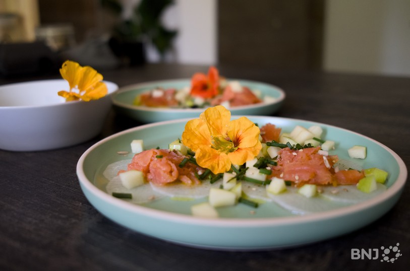 Une petite assiette avec du saumon posé sur des pétales de radis blanc et une fleur de capucine comestible…
